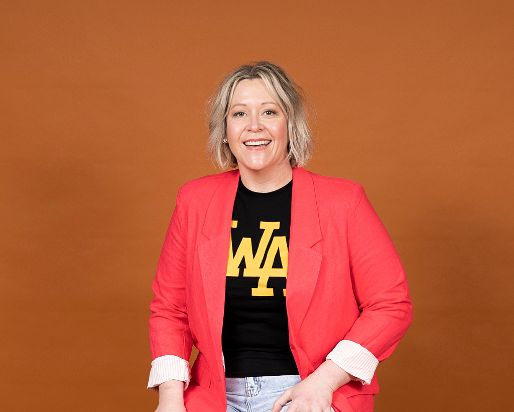 Andrea Gibbs smiling, a white woman with a blonde bob, wearing a black tshirt and red jacket