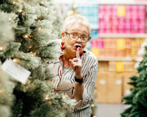 Sally-Anne Upton, a woman with short white hair, hiding behind a Christmas tree holding her finger to her mouth.