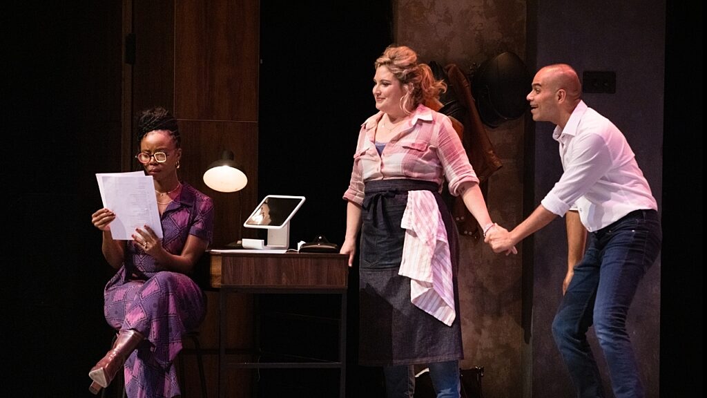 Two characters holding hands listening to a friend read a contract, on a stage in an office setting.