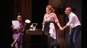 Two characters holding hands listening to a friend read a contract, on a stage in an office setting.