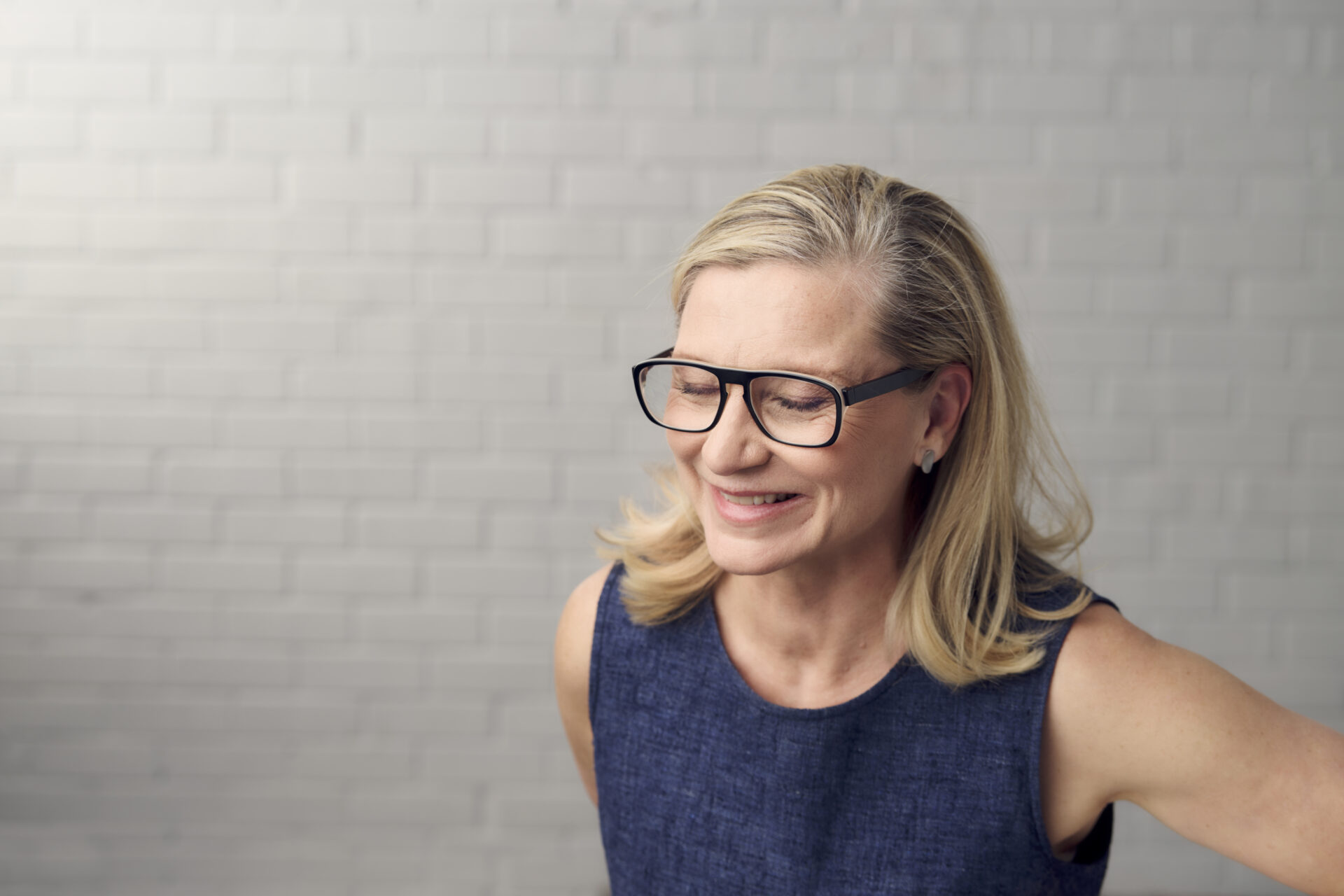 Kate Champion, a white woman with shoulder length blond hair smiling, wearing black glasses and a navy top against a white brick wall