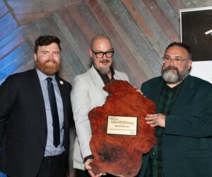 Man handing award to two other men standing against timebr wall