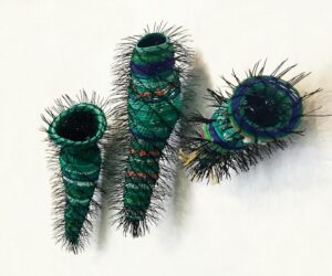 Three sculptural vessels made from layered recycled plastic in green and blue tones, wrapped with fine black spines, mounted on a white wall.