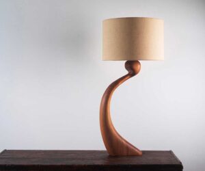 Curved timber table lamp by Margot Feast with a fabric lampshade on a dark wooden table.