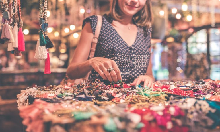 Woman not fully in frame at a markets looking through beads and bracelets on a table with other market stalls in the back ground out of focus