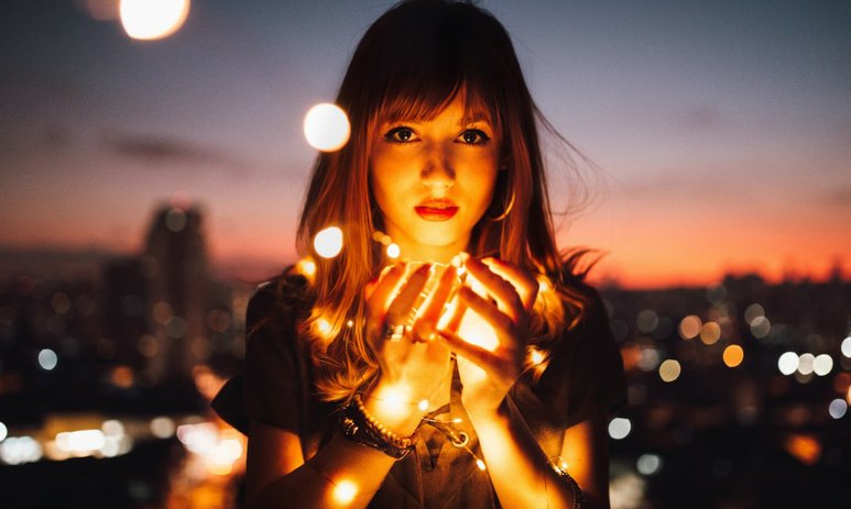 Young woman in focus holding warm string lights at dusk with skyline out of focus in background