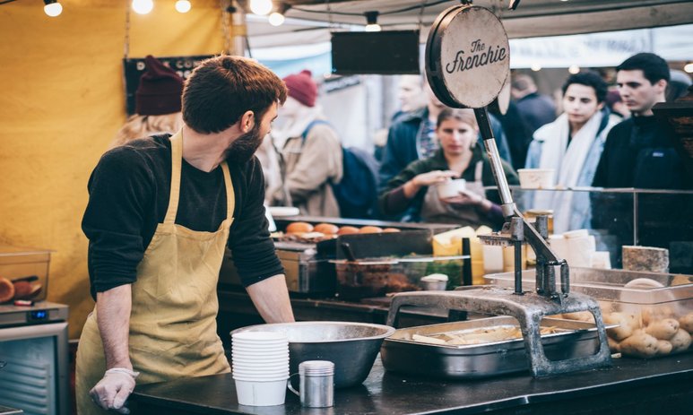 Market food vendor in his stall looking back at customers while leaning on food prep bench