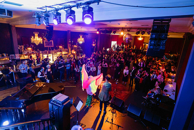 Large music venue with many people dresesd in ball attire listening to two people on stage; one in a top hat and one with inflated rainbow wings.