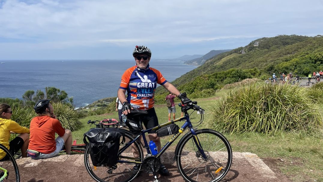 Cyclist standing with a bike at a scenic coastal overlook during the Great Cycle Challenge. Wearing cycling gear and a 'Great Challenge' jersey, with other participants and ocean views in the background.