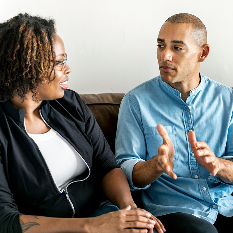 A couple sits side by side in therapy. The man gestures as he describes how he's feeling.