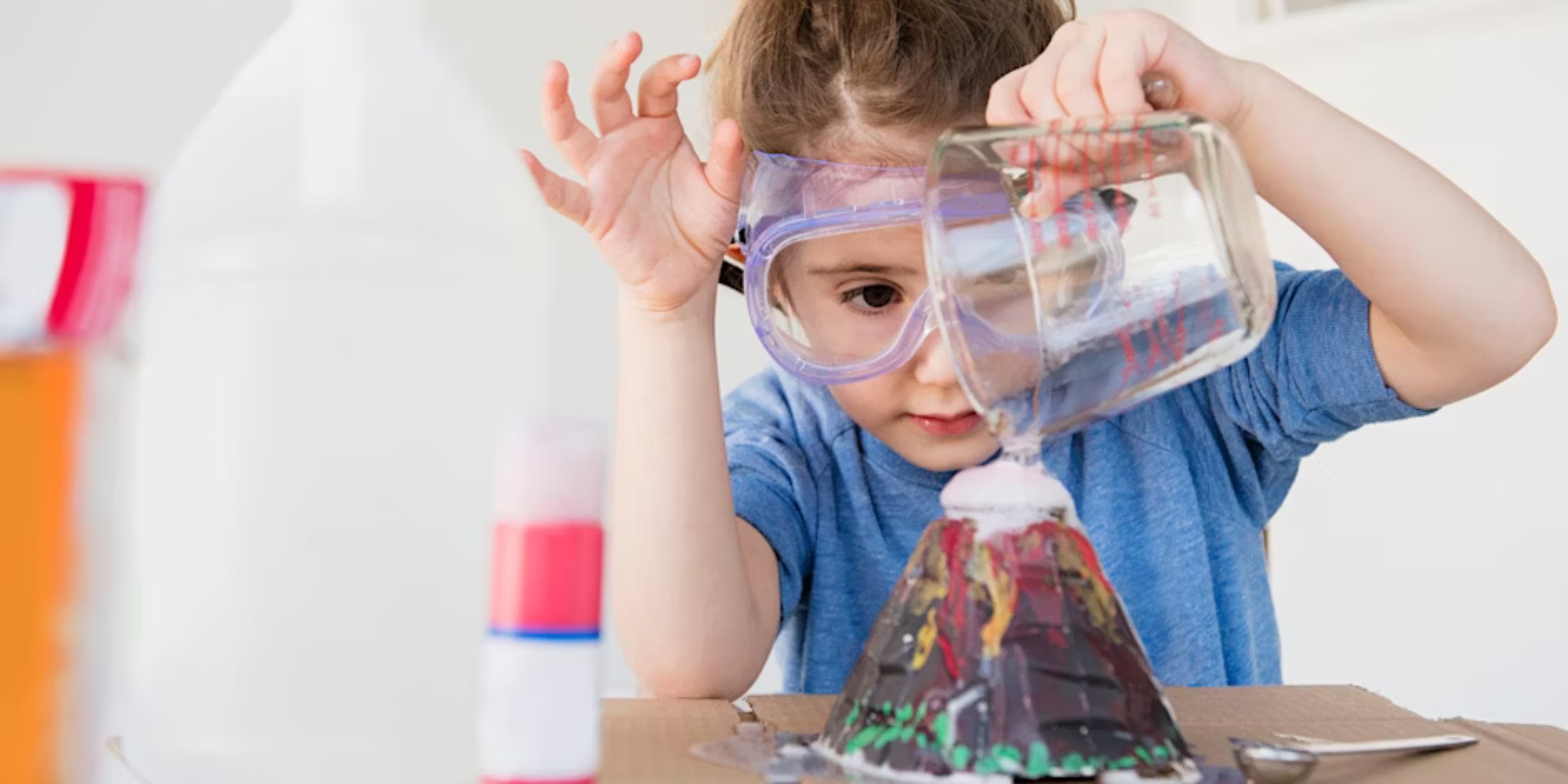 STEAM programs in Bunbury with Bunbury Public Libraries banner. The banner consists of an image of a children pauring a liquid into a volcano made of cardboard. The child is wearing safety glasses