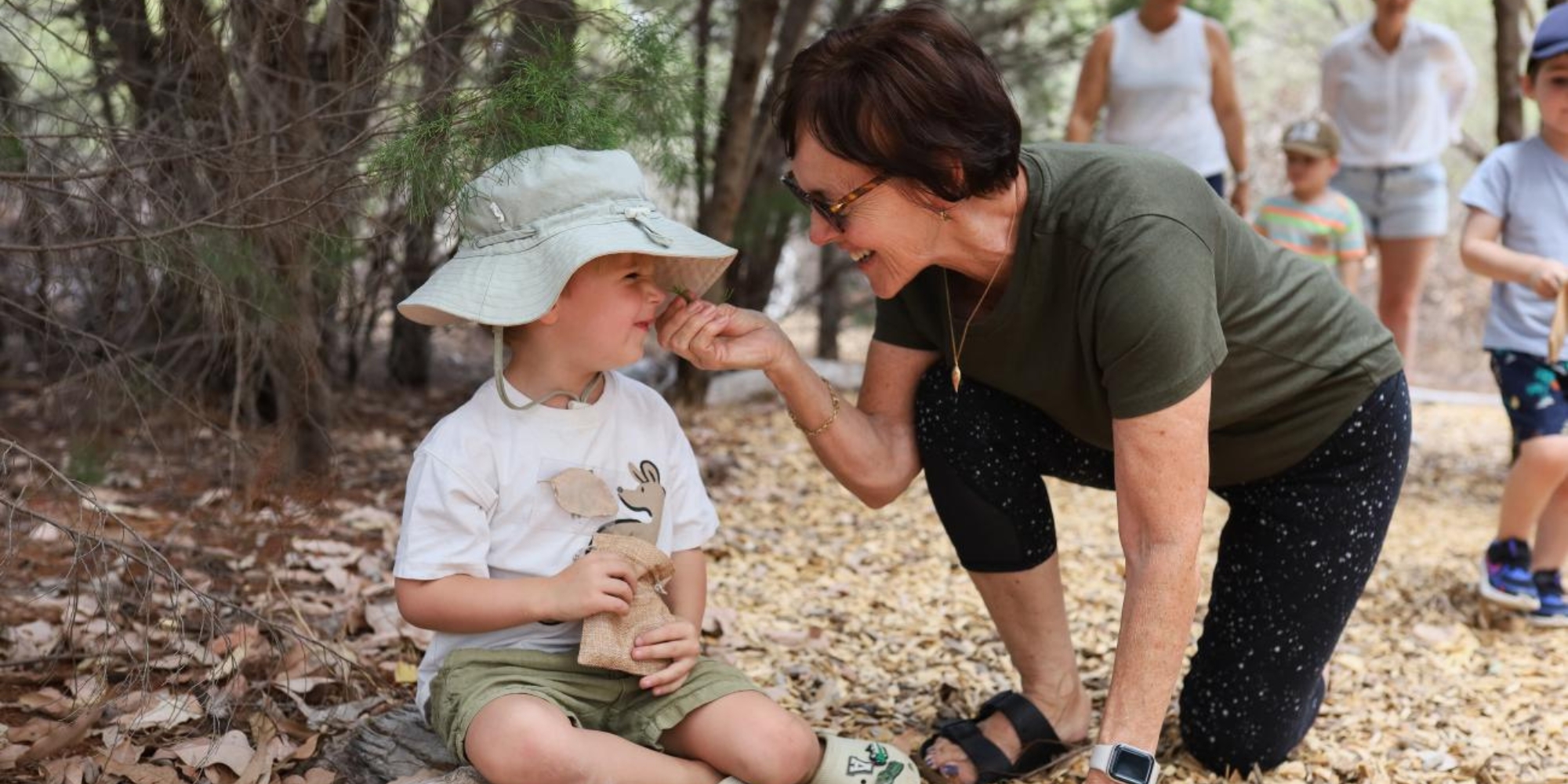 Zippys Bush Kindy with the Botanic Gardens and Parks Authority banner. The banner consists of an image of a child sitting down wearing a bucket hat. There is a woman kneeling on one knee and slightly touching the childs face. They are outdoors and surrounded by trees and dirt