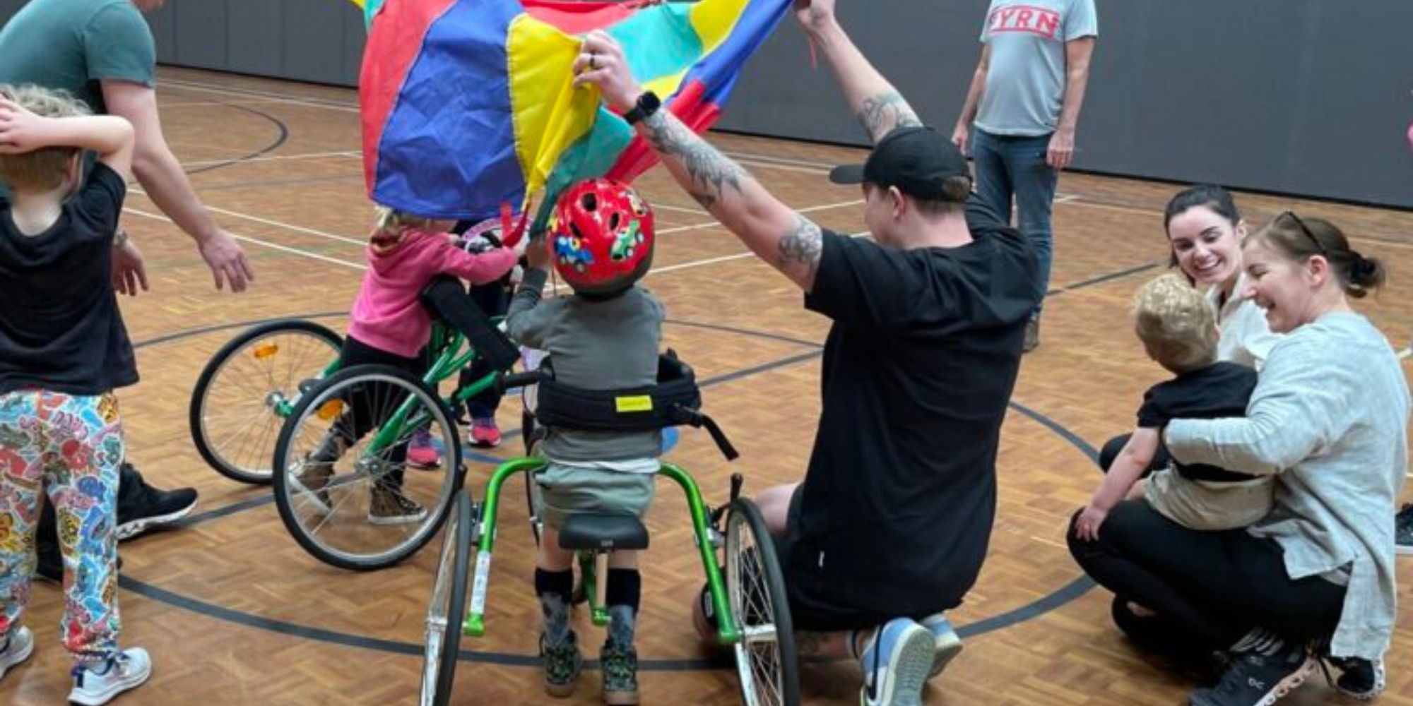 Run For Fun Frame Running Program with Rebound WA banner. The banner consists of an image of people on a indoor basketball court. There are children in FrameRunners and some family members and Rebound WA staff around the children laughing and smiling