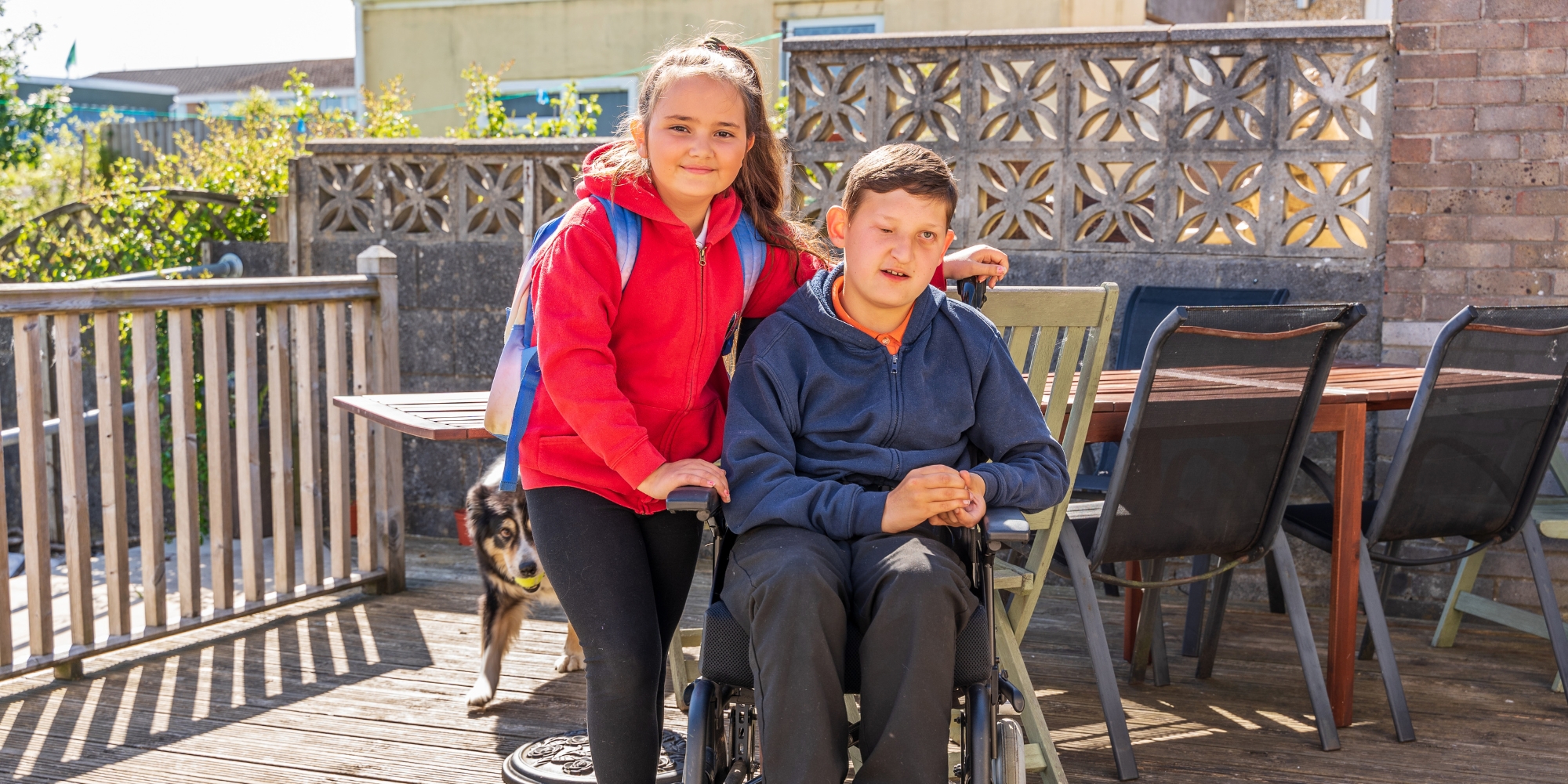 2026 Young Carer Bursary with the Young Carers Network banner. The banner consists of an image of a child and boy. The boy is sitting in a wheelchair and is wearing a navy jumper and the girl is standing next to him with her arms around his shoulders wearing a red jacket and backpack. They are both smiling at the camera.