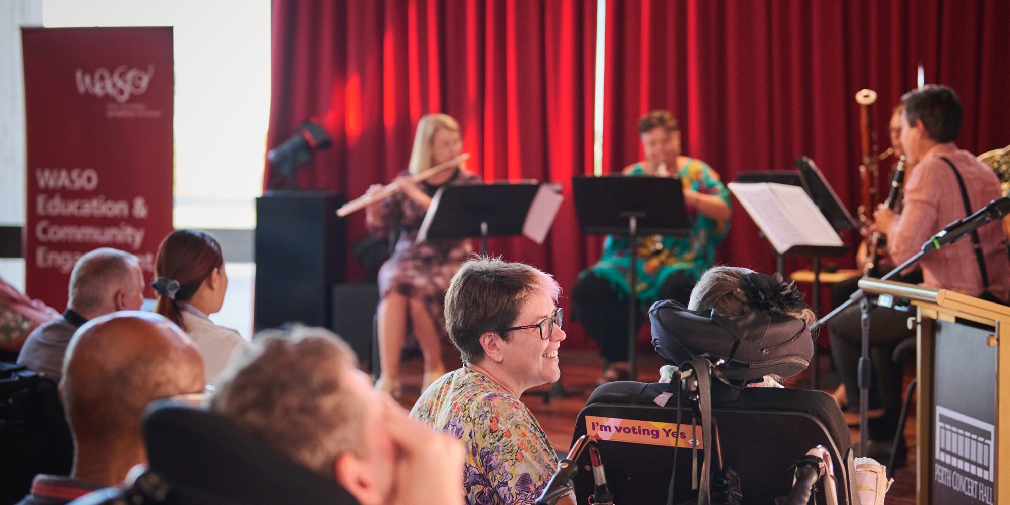 WASO accessible chamber concert with the West Australian Symphony Orchestra banner. The banner consists of an image of people in the audience and musicians on stage. There is a red curtin behind the musicans on stage.