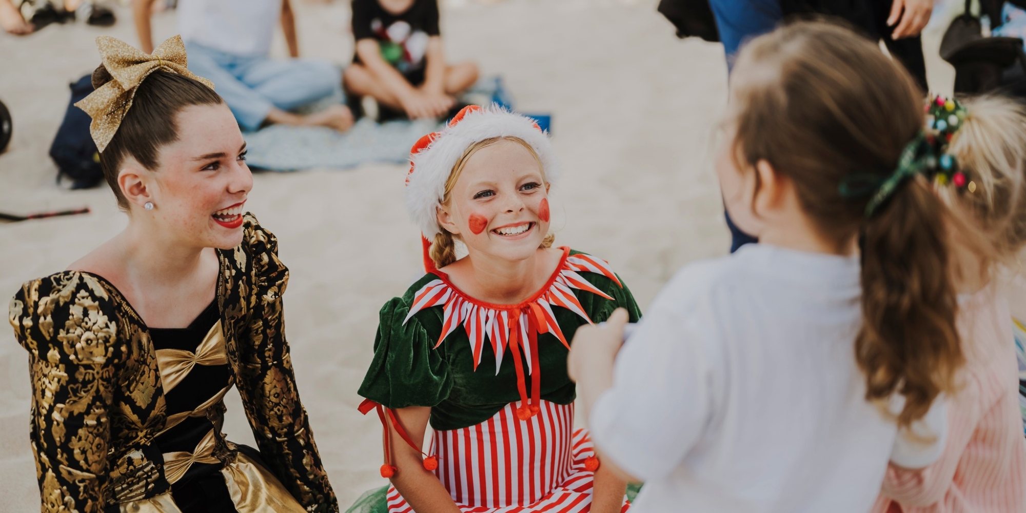 Carols at Scarborough with Neuromuscular WA and Scarborough Beach Association banner. The banner consists of an image of people dressed up on Christmas outfits while talking to children. The background is of sand on the ground at the beach.