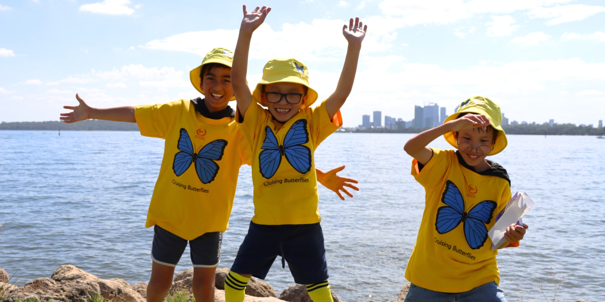 Cruising Butterflies 2026 with South of Perth Yacht Club, Rotary Club of Applecross and Melville and Kiind banner. The banner consists of an image of three boys who are wearing yellow Cruising Butterflies shirts and bucket hats. They are jumping up and cheering with the Swan River in the background.