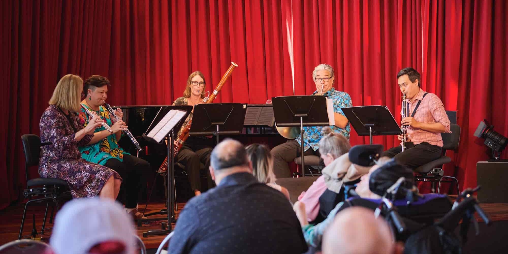 March Chamber Concert for all abilities with the West Australian Symphony Orchestra (WASO) banner. The banner consists of an image of wind musicians on a stage playing their instruments. There is a red curtain in the background with people watching in a crowd.