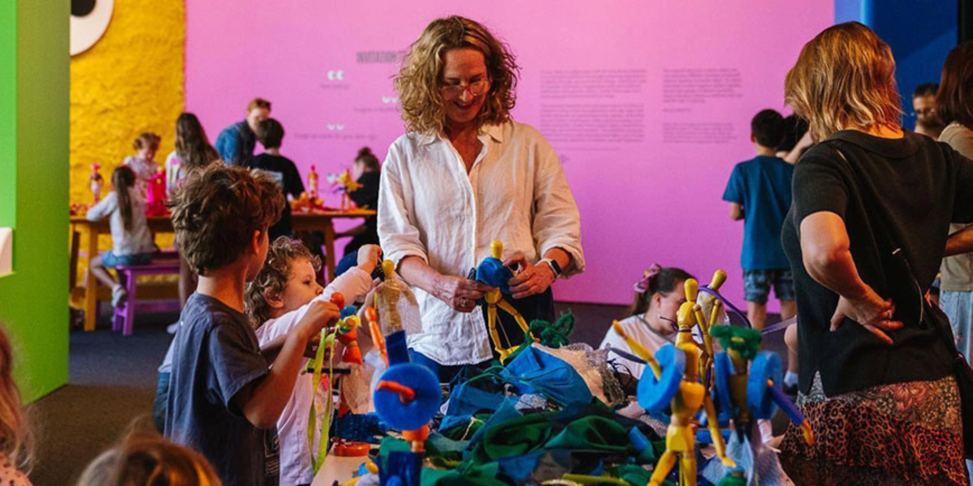 Cranky Pants Quiet Hour at the Art Gallery of WA (AGWA) banner. the banner consists of an image of a parent and children around a table of different coloured materials. The background is pink and yellow, blue and green. They are all smiling and interacting with the exhibition.