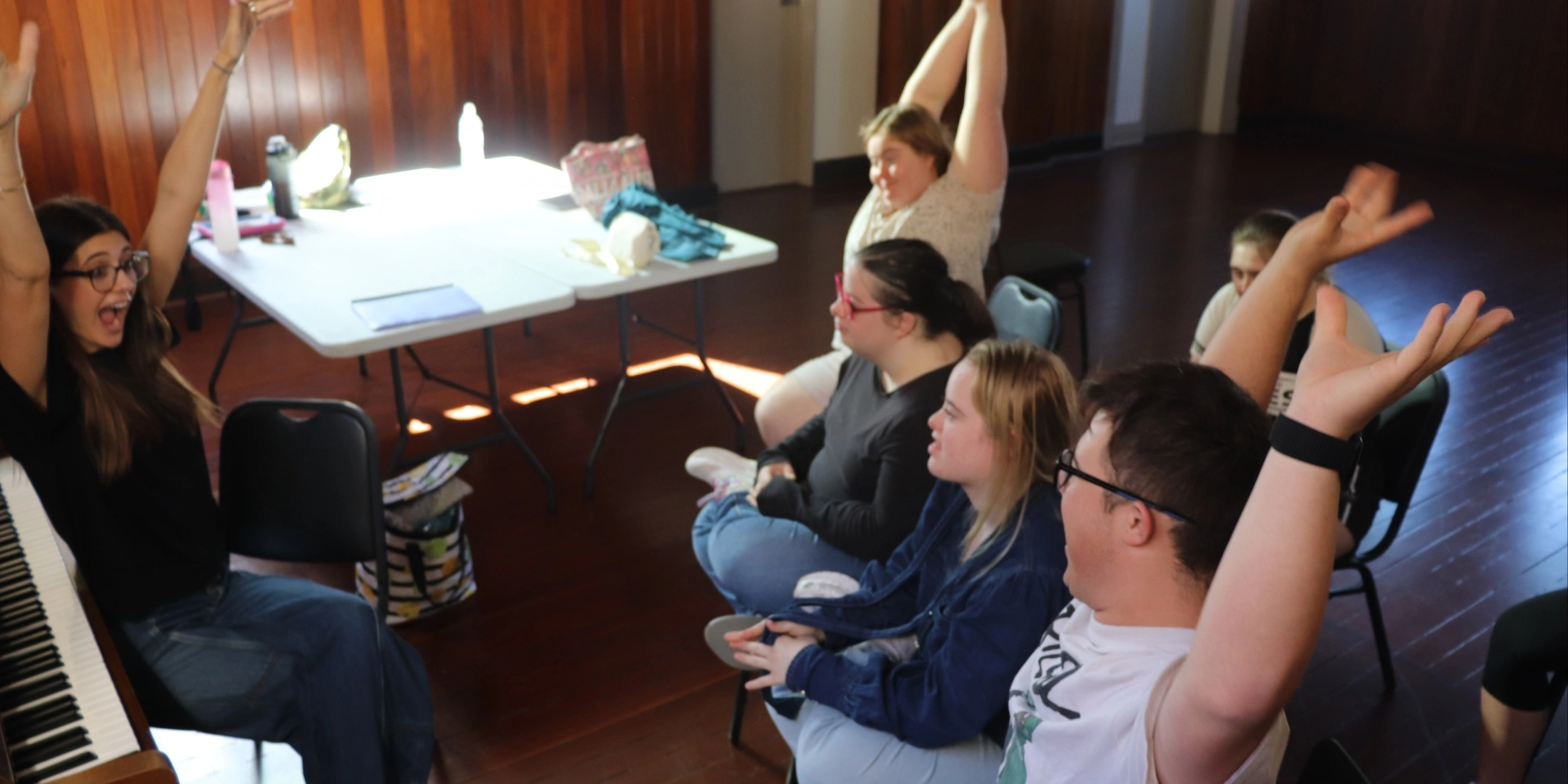 Acting course with Down Syndrome WA banner. The banner consists of an image of a groupd of people listen to an acting teacher with their arms up, just like the teacher. They are all sitting down in a room.
