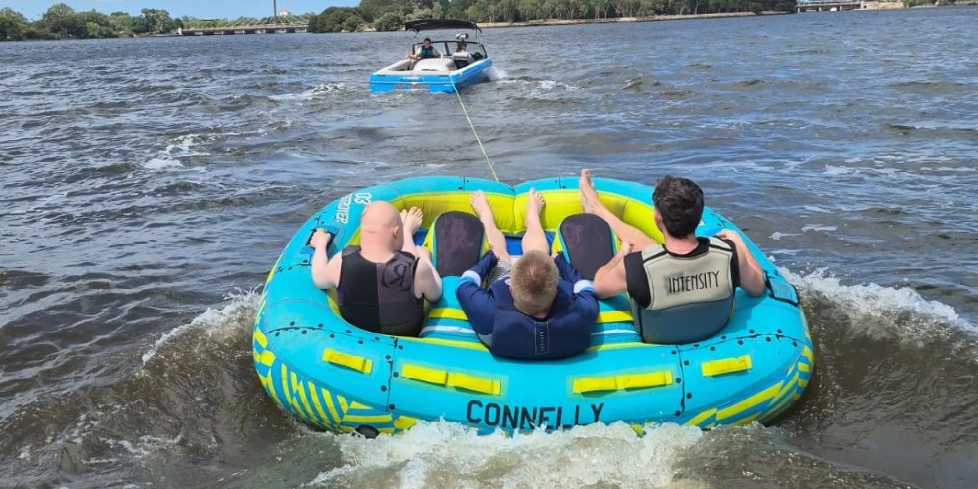 Three men sitting in an inflatable tube on the river with their backs to camera being towed by a boat