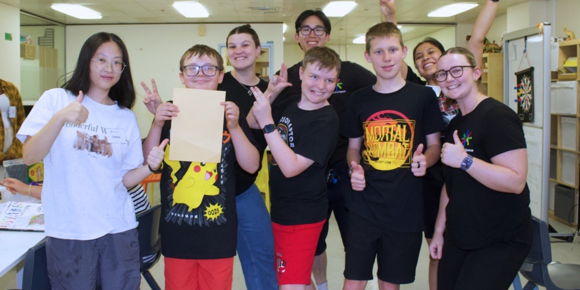 A group of teenagers smiling to the camera with some giving thumbs up and some giving peace signs