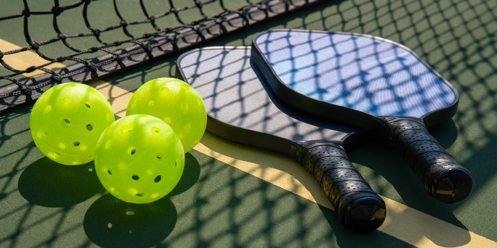 Three pickleballs and two pickleball paddles on a court next to the net