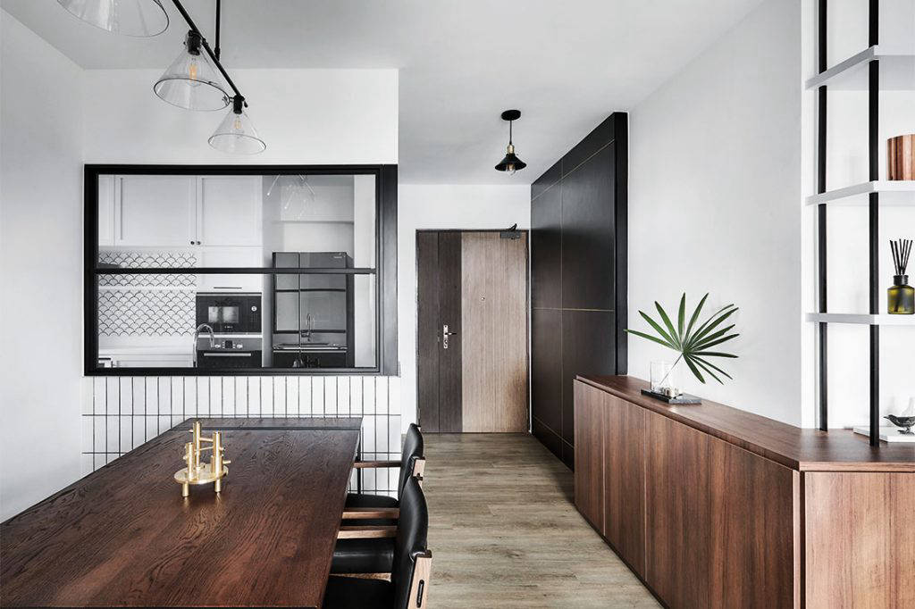 Form definitely follows function in this HDB flat for a young couple, where the UPSTAIRS_ design team has conceived a clean-lined yet edgy aesthetic. It shows in the pared-back setting of whitewashed walls, which highlights the custom carpentry in dark woodgrain accents. These clean forms of rich wood textures belie a generous amount of storage and are paired with black metal-framed shelving. Next to it is a black feature wall with brass trimmings that conceals the entrance to the household shelter. 