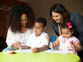 A mum and her two young children sit at a green table, colouring in with their therapist.