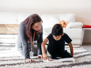 A young boy and his therapist sit on the carpet reading a book, with a white couch in the background.