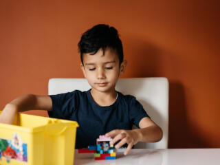 A child sits at a table with an orange wall behind him. He's taking some Lego out of a yellow plastic box.
