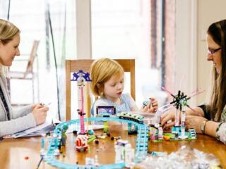 A blonde girl sits at a table with her mum and therapist, playing with building blocks.