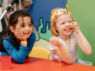 Two girls with party hats on sit smiling at a table with a colourful wall in the background.