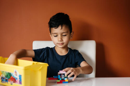 A child sits at a table with an orange wall behind him. He's taking some Lego out of a yellow plastic box.