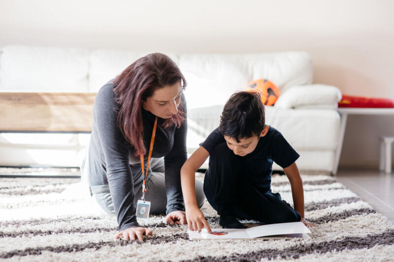 A young boy and his therapist sit on the carpet reading a book, with a white couch in the background.