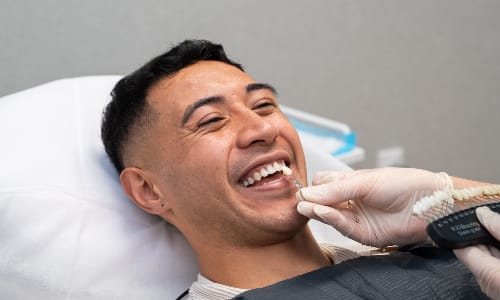 Male client sitting in a treatment chair while a therapist compares his teeth to a shade guide before whitening treatment.