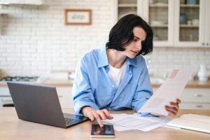 Woman sitting behind table at home kitchen, checking water bill, making calculations on calculator, paying utilities on laptop.