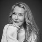 Black and white photograph of a woman with long hair wearing a cozy knit sweater, sitting with hands clasped near the face. The image focuses on texture and lighting, highlighting soft fabric details and hair strands against a plain dark background.