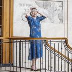 Photograph of a woman in a blue dress standing on an ornate staircase inside a library or historic building. She is smiling and holding a book on top of her head and there is , a marble wall with engraved text and a winged lion in the background.