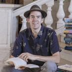 Photograph of a man in a hat sitting on marble stairs in a building, holding an open book with stacks of colorful books arranged on steps beside him. The setting includes white balusters and a plaque on the wall, suggesting a library or academic environment.