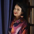 Photograph of a woman partially hidden behind dark blue curtains, with only their upper body visible. The person wears a colorful, patterned shirt with shades of pink, purple, and orange, standing against a dimly lit background with wooden elements.