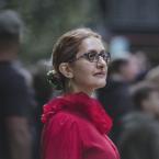 Photograph of a woman wearing glasses and a bright red blouse with ruffled collar standing outdoors among a blurred crowd. Focus is on the upper body and profile, highlighting a green hair accessory and dangling earrings against a soft, natural background.