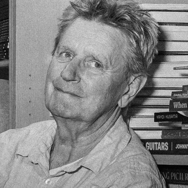 Photograph of a man sitting on a box in a cluttered storage room filled with shelves of books and stacked cardboard boxes. The person is casually posed with arms crossed, surrounded by various items including a trolley and scattered books on the floor, suggesting a setting related to book storage or inventory.