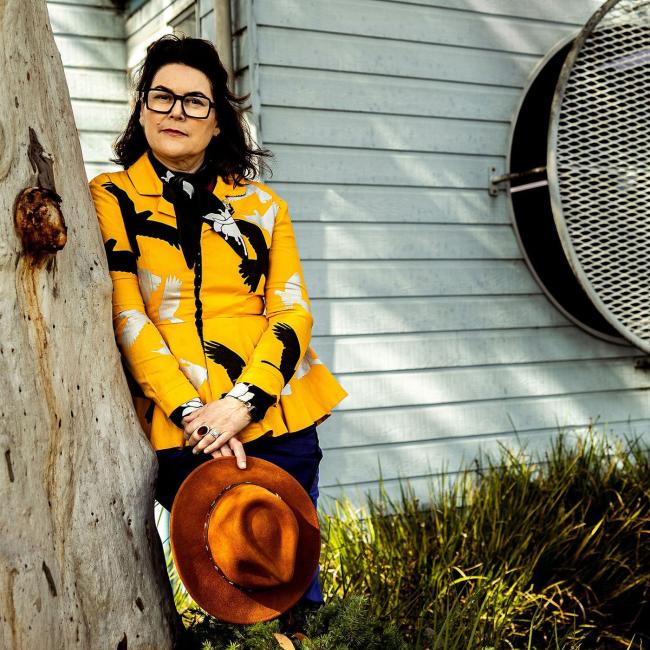 Photograph of a woman outdoors leaning against a tree in front of a small light blue wooden structure with a large circular vent. The person wears a yellow patterned jacket and holds a brown hat, with greenery and sunlight creating a natural, serene setting.