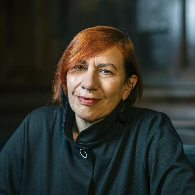 Photograph of a smiling woman with short red hair sitting indoors, wearing a dark jacket and a white shirt underneath. Background features dim lighting and dark wooden panels, suggesting a formal or professional setting.