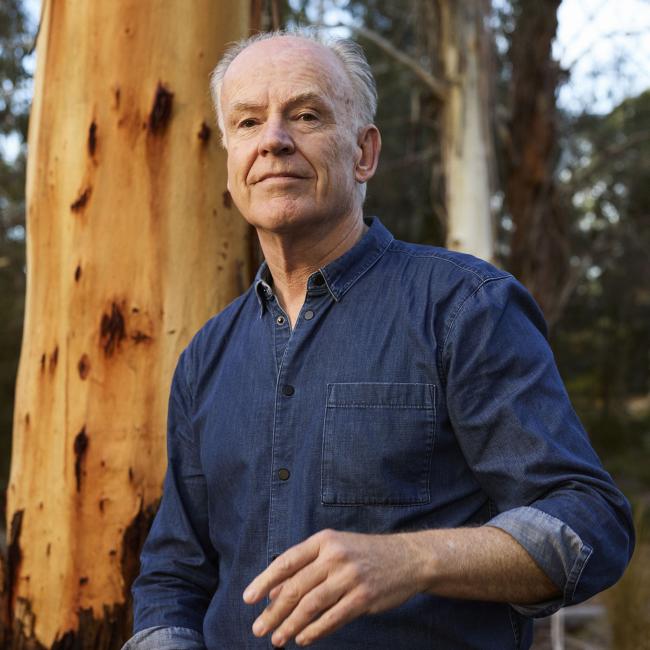 Photograph of a man standing outdoors in a wooded area with tall trees, wearing a blue denim shirt and dark jeans. The man is positioned slightly off-center, with one hand raised and the other holding a hat, suggesting a casual or conversational moment.