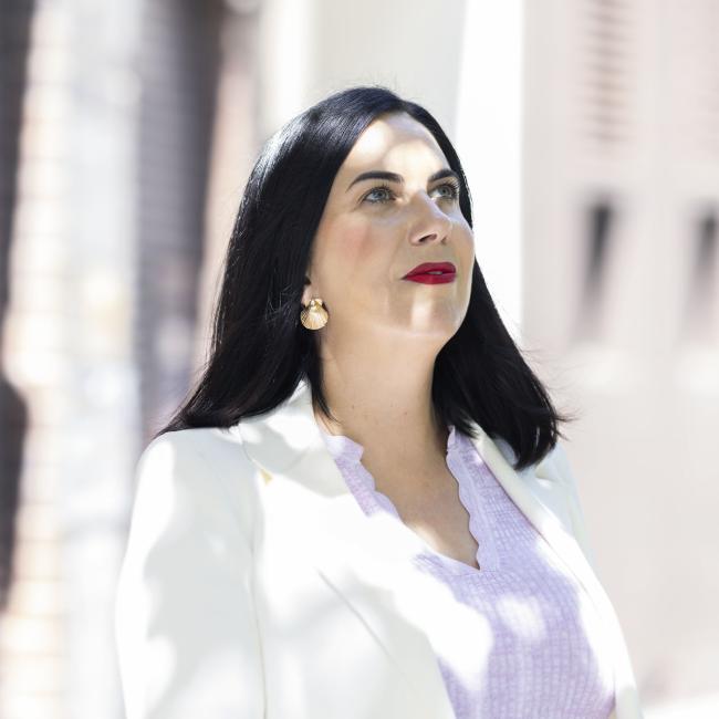 Photograph of a woman with long black hair wearing a white blazer and light purple top standing outdoors against a blurred brick wall background. Her face is turned upwards, with soft dappled natural light on her face.