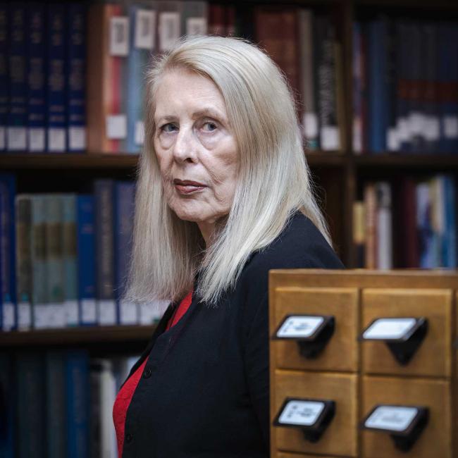 Portrait photograph of an older woman with long silver blonde hair standing beside wooden card-catalog drawers in a library or archive. Bookshelves and labeled drawers create an academic research setting, while subject’s direct gaze forms primary visual focus.
