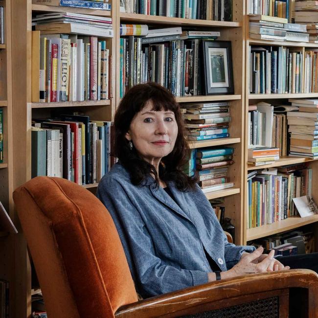 Photograph portraying a dark haired woman seated in a burnt orange armchair, surrounded by floor-to-ceiling bookshelves in a home library.
