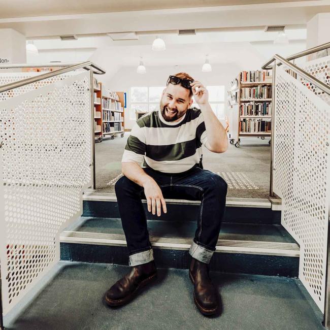 Photograph of bearded man seated on library stairs, wearing striped shirt, cuffed jeans, boots, and sunglasses on his head. Bright library setting features bookshelves, white perforated railings, pendant lights, and windows, creating a casual portrait scene.